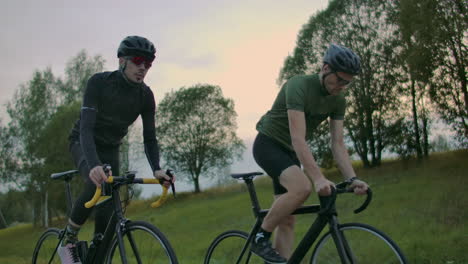 Gutaussehender-Bärtiger-Professioneller-Radfahrer,-Der-Morgens-Zusammen-Mit-Seiner-Freundin-Auf-Seinem-Rennrad-Fährt,-Beide-Tragen-Schutzhelme-Und-Brillen,-Zwischen-Denen-Die-Sonne-Durchscheint